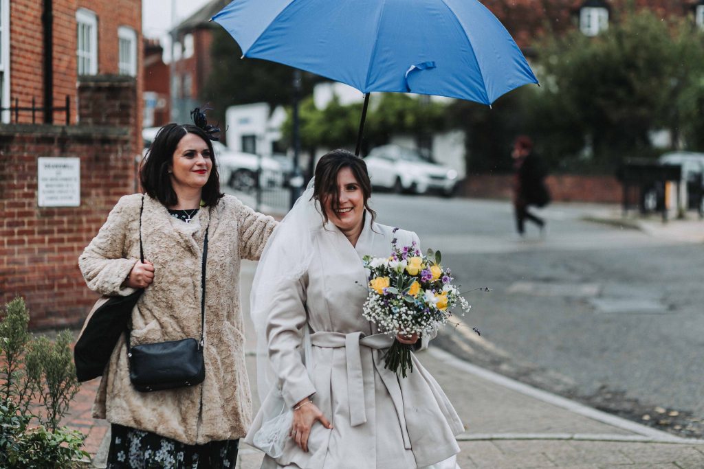 bride with bridesmaid walking with umbrella in rain laughing