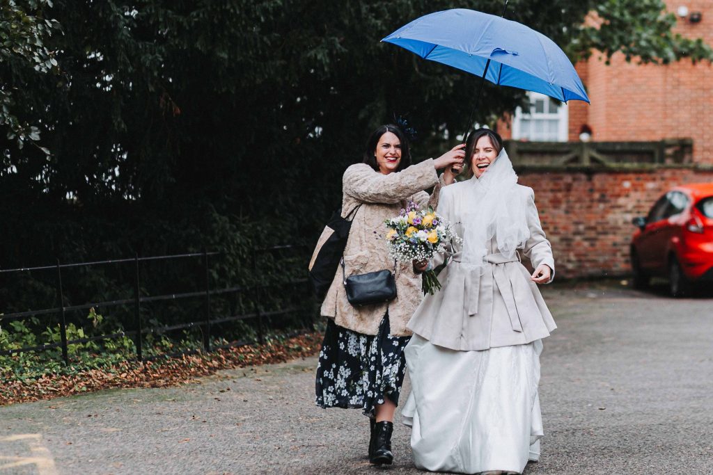 bride with bridesmaid walking with umbrella in rain laughing