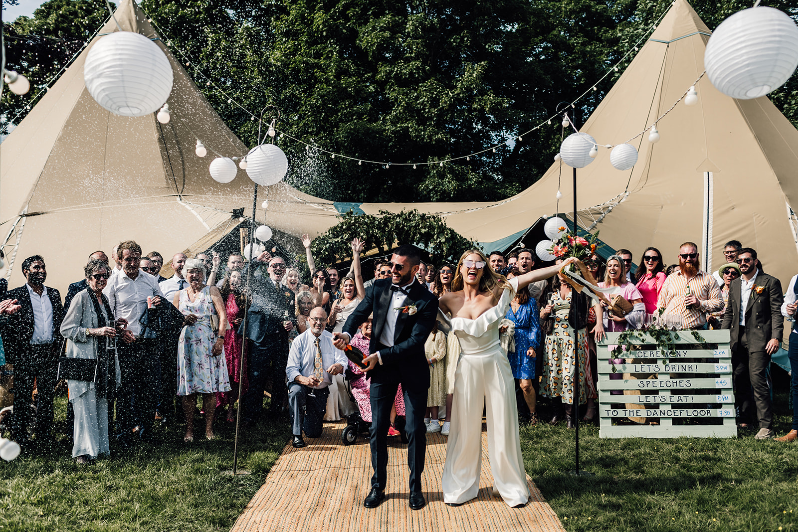 Bride and Groom popping champagne in front of wedding guests at tipi festival styled wedding.