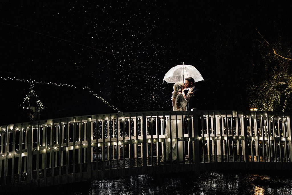 couple portrait on a white bridge over a small pond at night in rain with umbrella at Winters Barns wedding venue