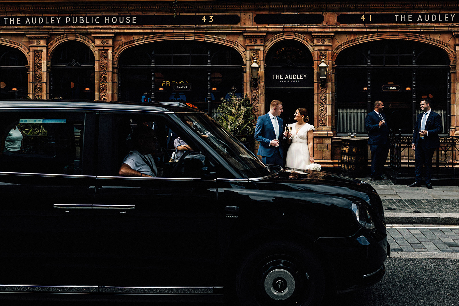 bride and groom documentary portrait on streets of Mayfair, London holding glasses of champagne outside The Audley pub with black taxi cab driving past.