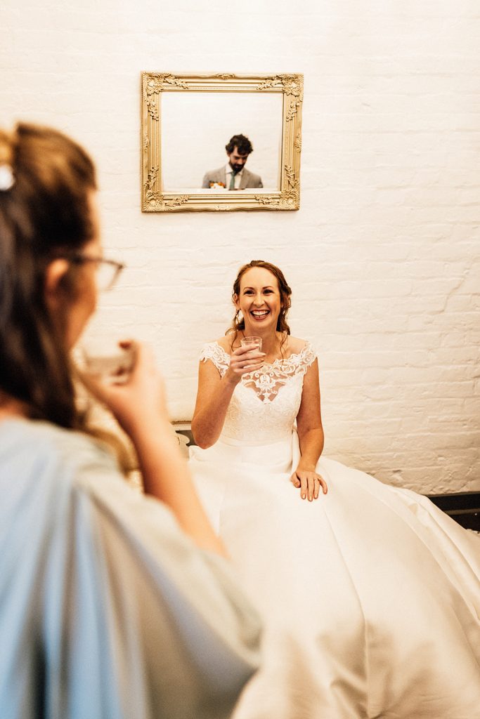 bride in wedding dress sitting down holding a drink laughing towards camera with friend looking on