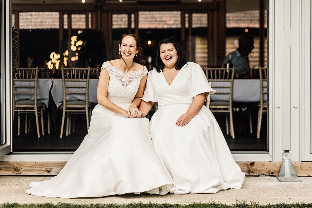 candid portrait of 2 brides in white wedding dresses sitting down laughing and smiling