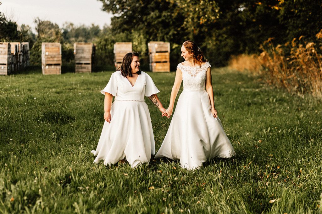 2 brides holding hands in white wedding dresses in field looking at each other and smiling.