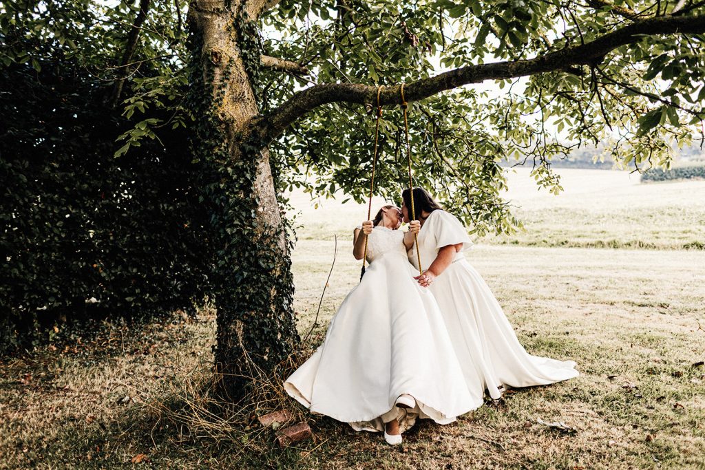 2 brides in white dresses by a tree swing in the grounds of The Night Yard wedding venue