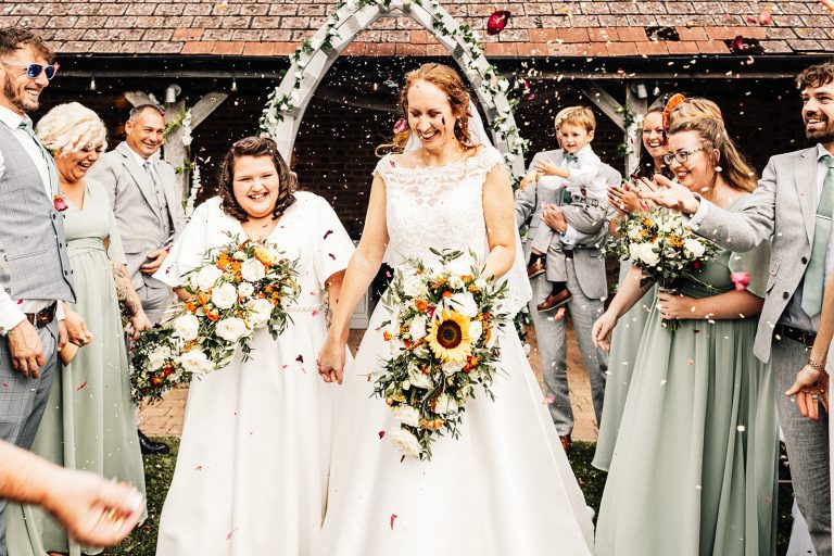 2 brides smiling walking down the aisle in an outside ceremony being showered with confetti by their guests.
