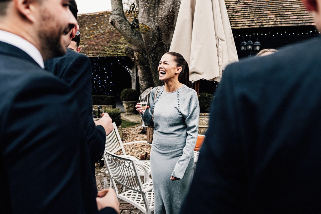 female wedding guest holding a glass of prosecco laughing in company.