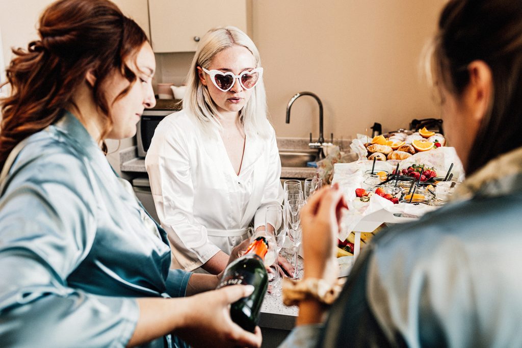 bride with heart shaped glasses on and in a dressing gown being poured a glass of prosecco while getting ready at bridal prep.