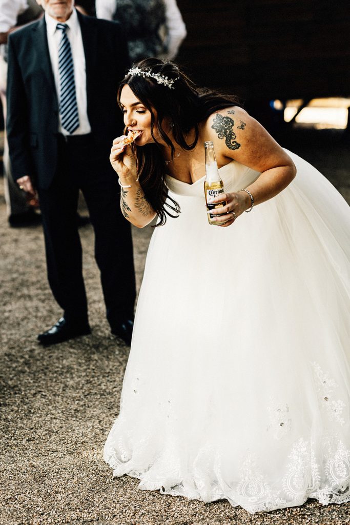 bride bent over eating pizza so as not to ruin her dress while holding a beer capyured by Chilham Village Hall wedding photographer