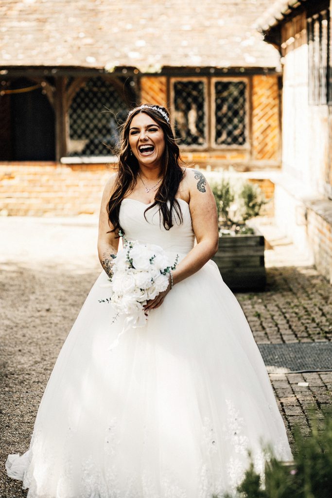 Chilham Village Hall wedding photographer captures bride dressed in sleeveless wedding dress holding bouquet of flowers laughing