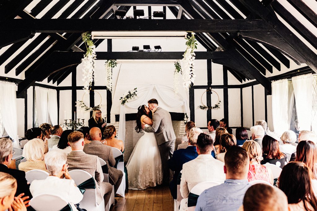 newly married bride and groom kissing in front of guests at a Chilham Village Hall wedding