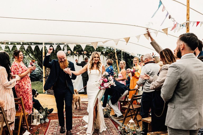 bride and groom just married exit in tipi wedding with them holding hands and groom punching the air walking down aisle past guests.