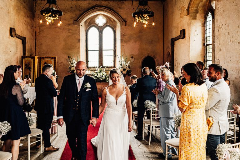 bilsington priory just married exit walk from ceremony by bride in white dress and groom in dark suit with pink tie