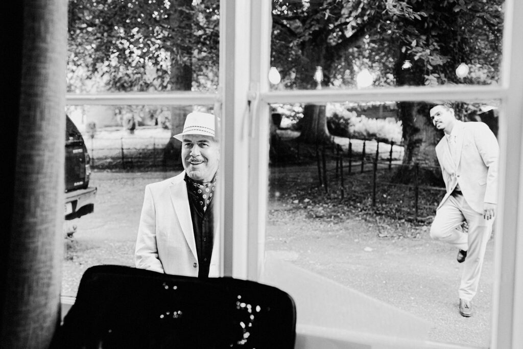 Father of bride dressed in suit and fedora hat looking in through the window of where the bride is getting ready at Swarling manor wedding venue.
