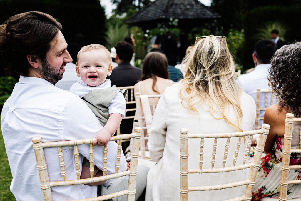 baby being held by his dad looking towards camera and laughing at an outdoor wedding ceremony at Swarling Manor wedding venue.