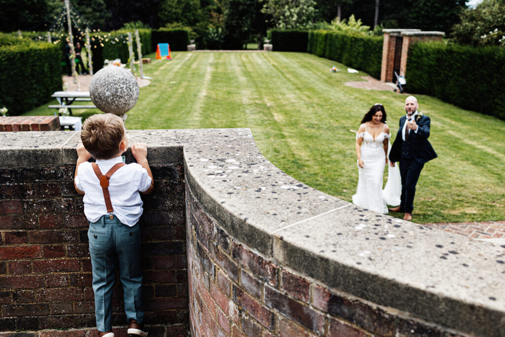 young boy peeking over a wall spying on his mum and dad who are the bride and groom in the gardens of Swarling manor wedding venue.