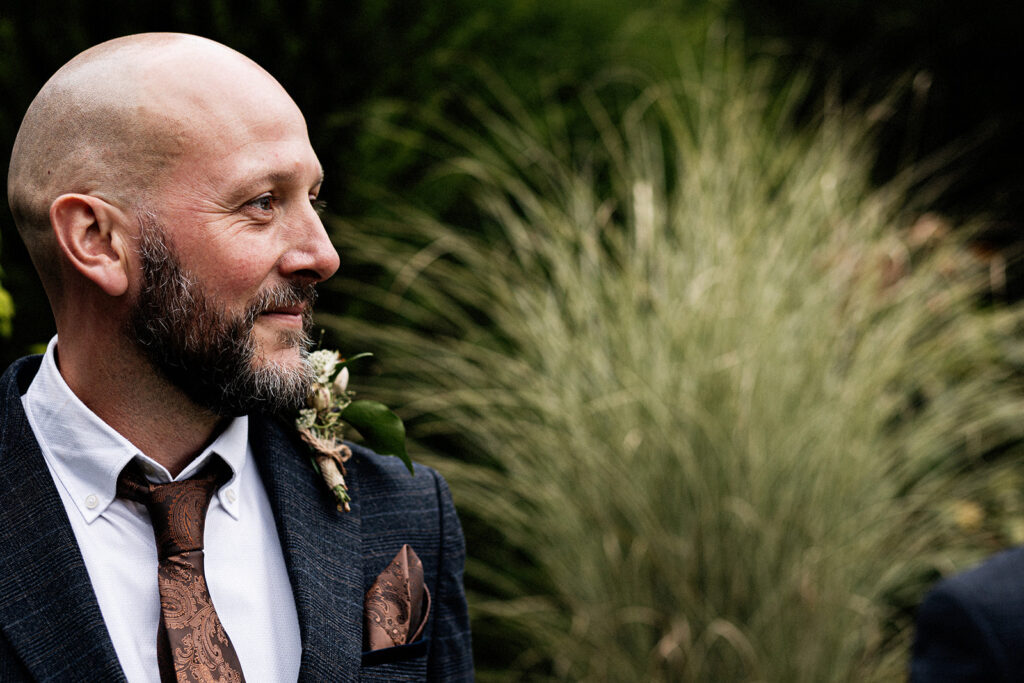close up profile picture of groom in blue suit, white shirt and bronze tie looking towards his bride who is out of camera frame walking towards him down the aisle at an outdoor wedding ceremony at Swarling manor wedding venue.