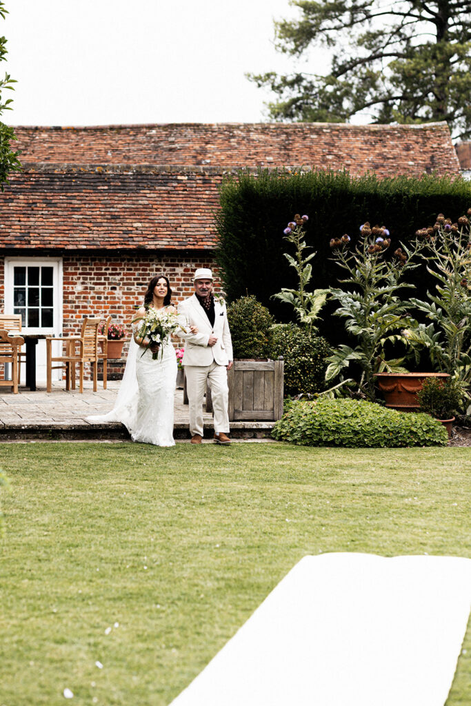 Father of the bride dressed in white suit and white fedora hat walking the bride towards the aisle who is also dressed in white holding flower bouquet towards the outdoor ceremony at Swarling Manor wedding venue.