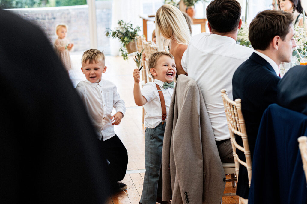 young boys playing around during wedding breakfast poking guests with leaves from the table displays in the marquee at a Swarling manor wedding.