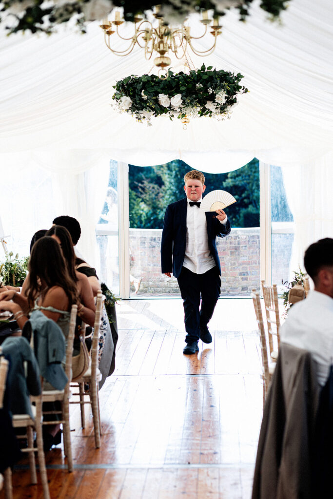 a boy in a tuxedo at a wedding walking down the marquee at Swarling manor wedding venue using a paper fan on a hot day.