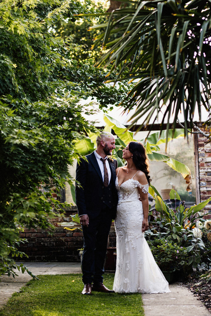 bride and groom portrait standing smiling at each other on a pathway between trees and large plants at Swarling Manor wedding venue.