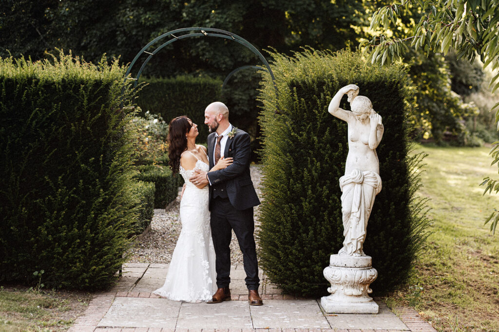 wedding portrait of bride and groom holding each other and looking at each other under an archway and next to tall hedges and a white marble statue in the grounds of Swarling manor wedding venue.